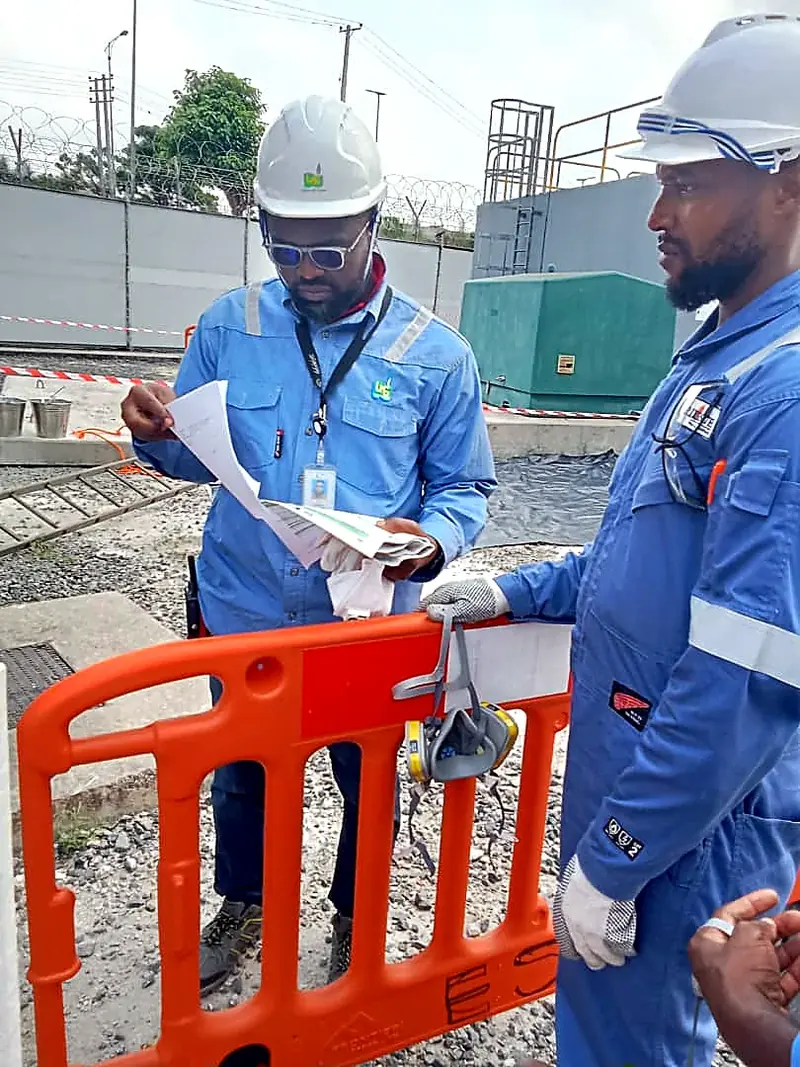 Two colleagues in blue coveralls reviewing paperwork beside orange barriers